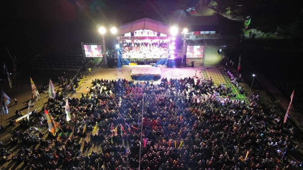 Foto : Pemandangan udara atau wide shot Lapangan Tembak Kodim 0812 Lamongan yang diterangi cahaya, dipadati ribuan orang saat acara sholawat akbar.
